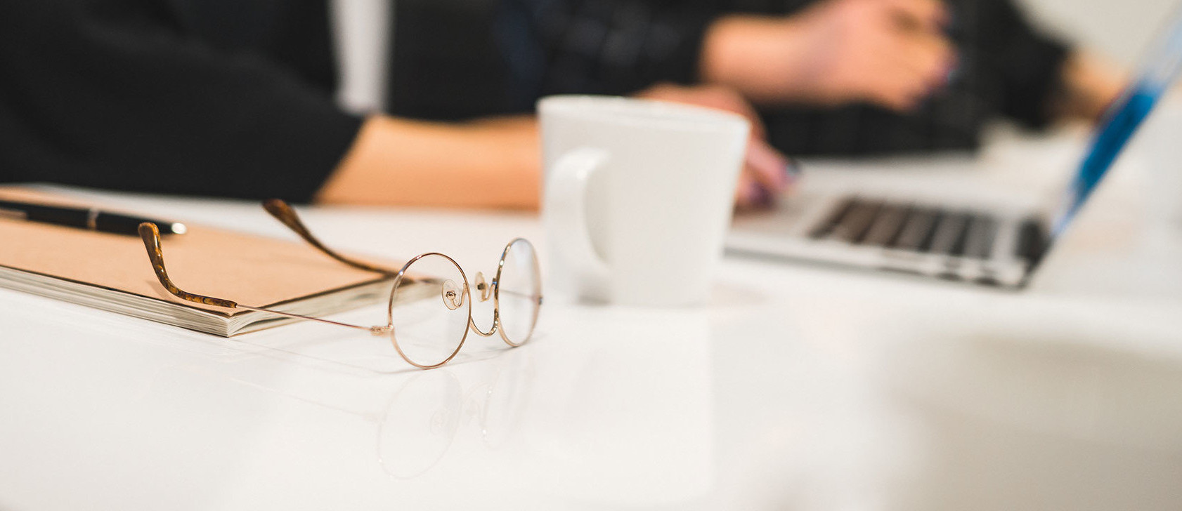 Ett par runda glasögon ligger på ett bord. I bakgrunden arbetar två personer på sina datorer.